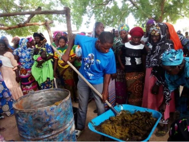 Homme malien en démonstration à Sitantoumbou. Il prend du compost dans une brouette à l'aide d'une pelle et s'apprête à le déposer dans le baril au premier plan. Il est entouré d'un groupe de personnes attentives. 
